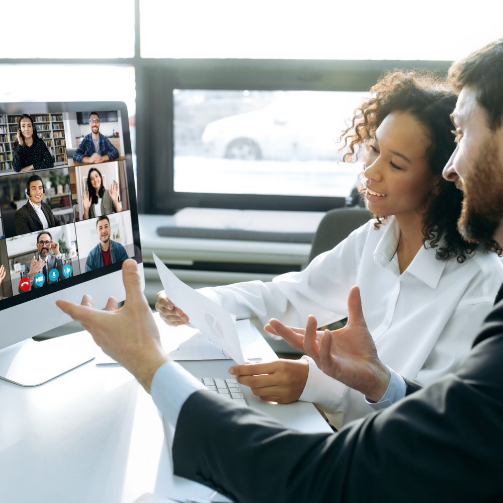 Business colleagues, co-founders of the company, sit at a table in the modern office in front of a computer screen, hold an online video conference with international colleagues, brainstorming