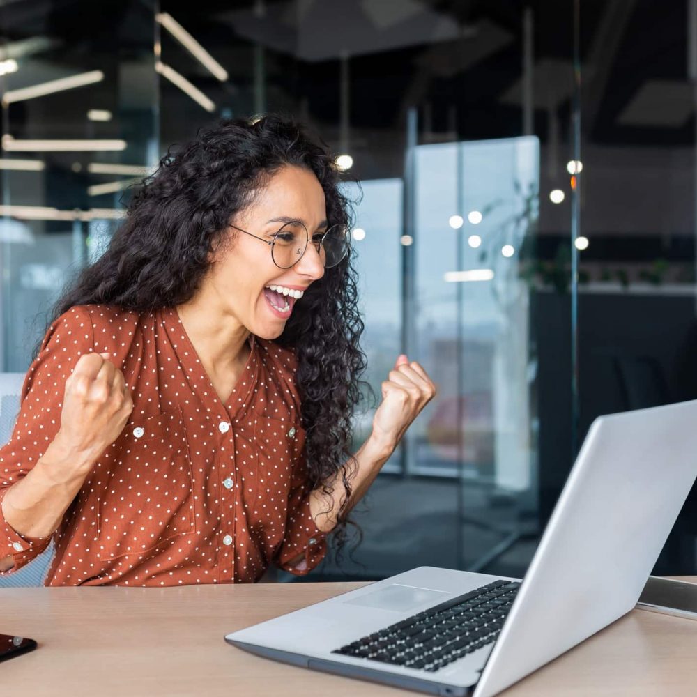 Hispanic business woman celebrating victory success, employee with curly hair inside office reading good news, using laptop at work inside office holding hand up and happy triumph gesture.